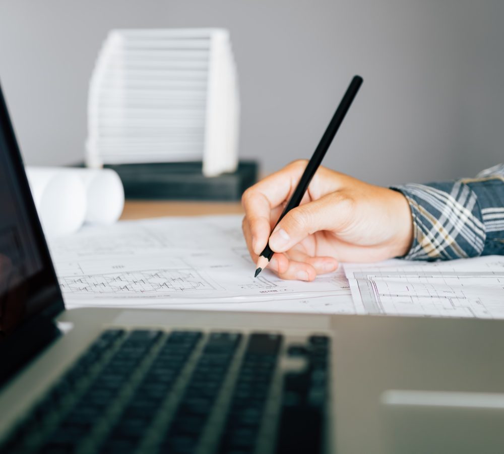 Hand of young engineering man drawing on blueprint with laptop on desk.